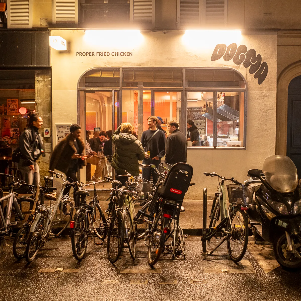 Evening outside a lit restaurant, bikes and scooters parked in front of the crowd.